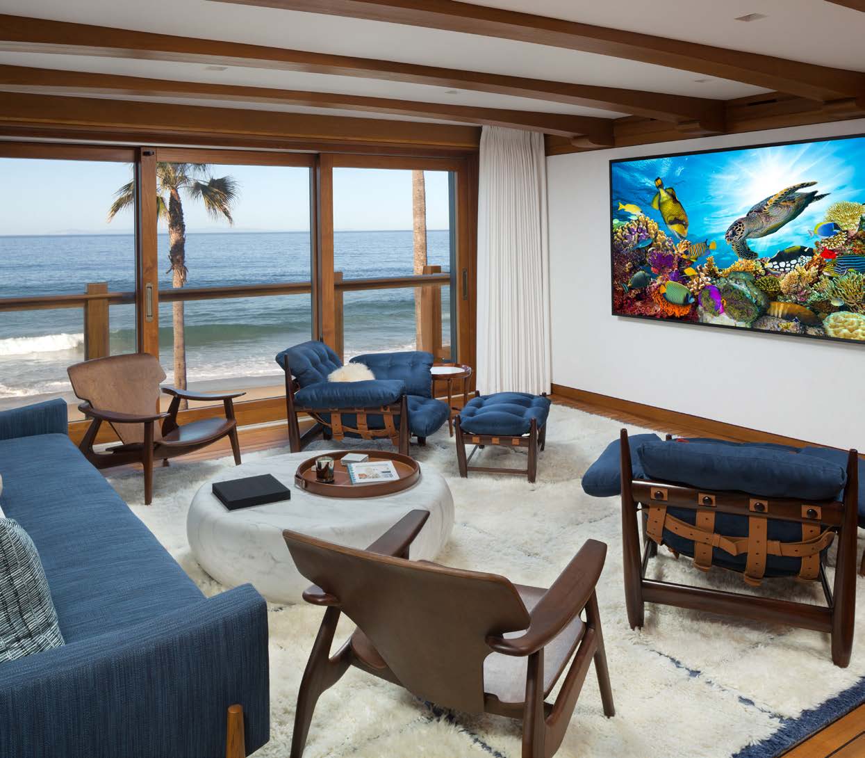 The finished media room — mid-century blue seating on a white rug, floor-to-ceiling glass to the Pacific on the left, and the concealed 98-inch 4K display on the right wall.